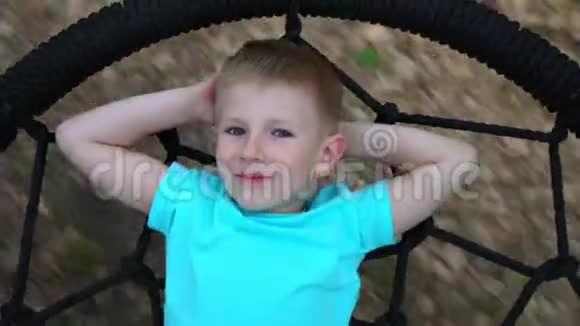 那个蓝眼睛的五岁男孩躺在一个圆秋千上双手放在头后看着相机微笑着视频的预览图
