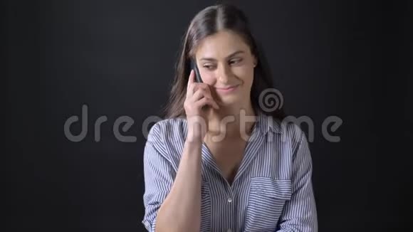 迷人的快乐的年轻女子一边打电话一边微笑独自站在黑色的背景下视频的预览图