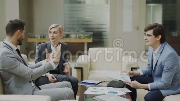 商人与坐在现代办公大厅沙发上的男女商业伙伴讨论财务报告视频的预览图