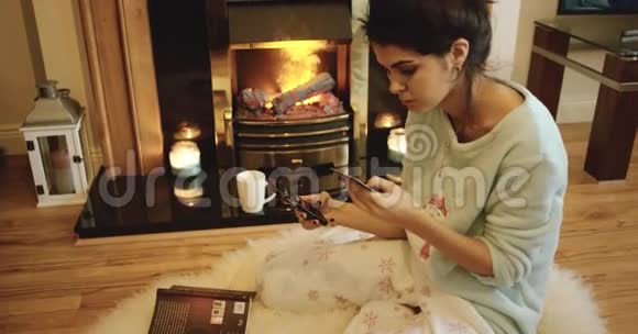 一个迷人的女人坐在炉火旁用信用卡从她的手机上订购视频的预览图