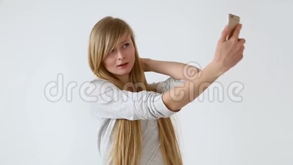 现代青年漂亮的长头发的欧洲女孩金发在靠近白色墙壁的智能手机上自拍视频的预览图
