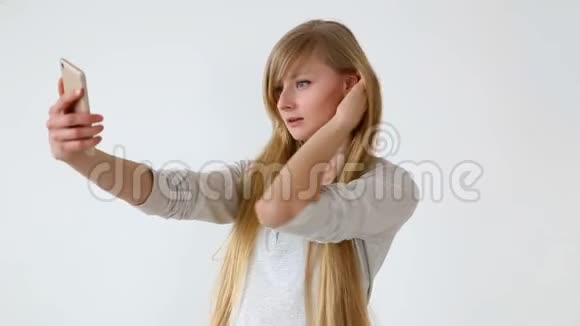 现代青年漂亮的长头发的欧洲女孩金发在靠近白色墙壁的智能手机上自拍视频的预览图