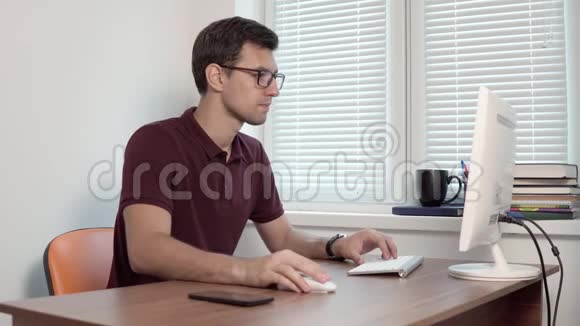 年轻的办公室工作人员戴着眼镜坐在窗边的办公室里用电脑打字报告智能手机视频的预览图
