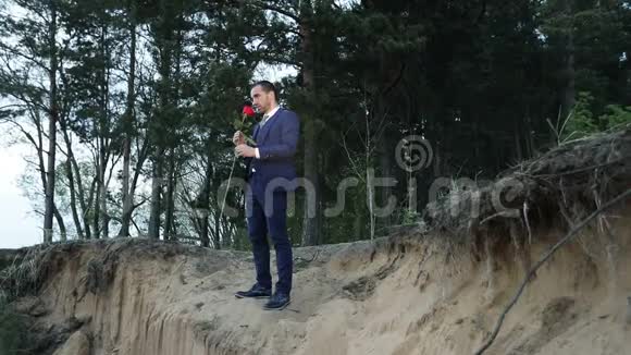 一个悲伤的商人站在湖边的悬崖上把一朵红玫瑰扔进水里视频的预览图