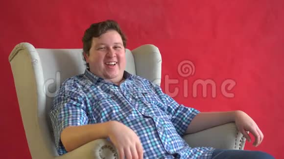 年轻的胖子穿着格子衬衫坐在椅子上在红色背景下大声笑着有趣的概念视频的预览图