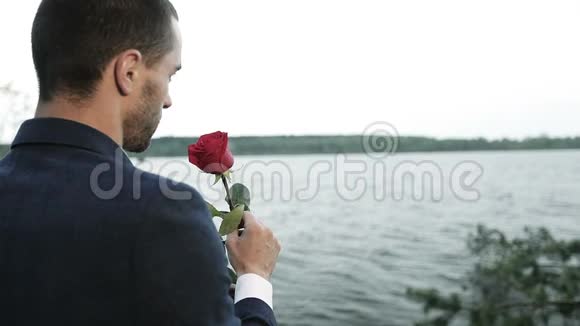 一个穿西装的年轻商人手里拿着一朵红玫瑰在湖边视频的预览图