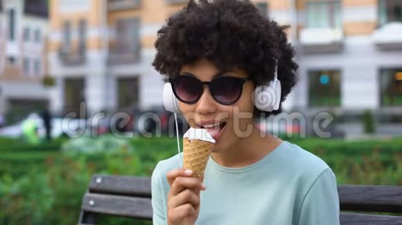 富有创意的无忧无虑的女人戴着耳机欣赏音乐在长凳上吃冰淇淋视频的预览图