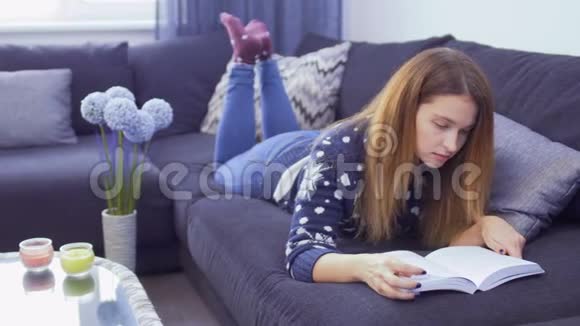 年轻可爱的女孩在家里的沙发上轻松地阅读这本书视频的预览图