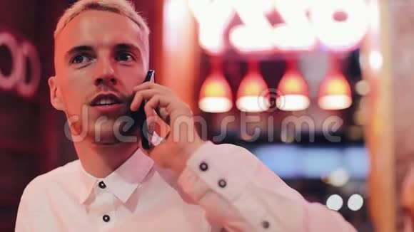 年轻的帅哥在夜间咖啡馆的智能手机上聊天他坐在霓虹灯招牌附近通信和业务视频的预览图