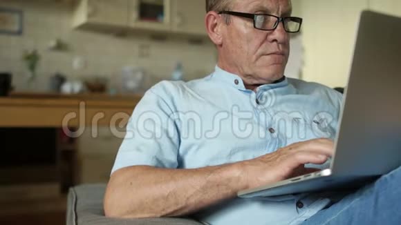 老人坐在厨房里用笔记本电脑工作他放松静静地微笑一个穿蓝色衣服的男人视频的预览图