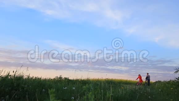 浪漫的散步两个可爱的恋人骑着自行车在美丽的绿色田野上美丽的天空视频的预览图
