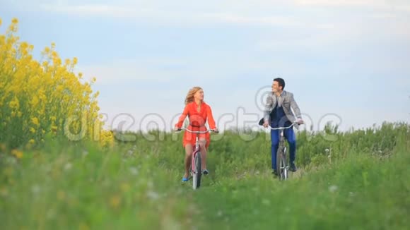 正面看着美丽欢快的情侣们积极骑着自行车沿着绿色盛开的田野视频的预览图