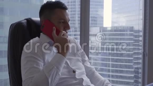 穿着白色衬衫的漂亮黑发男子在现代办公室的智能手机上讲话视频的预览图