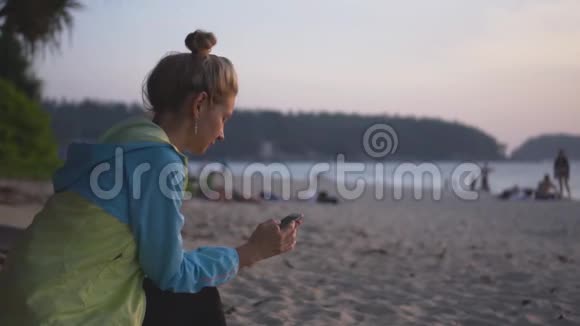 日落时分带着电话的悲伤女孩坐在海滩上视频的预览图