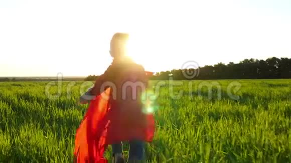 一个穿着超级英雄服装的孩子穿着红色斗篷在日落的背景下穿过绿色的草坪那个男孩视频的预览图