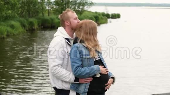 一位孕妇在湖边拥抱她的丈夫年轻的家庭期待着孩子走进来视频的预览图