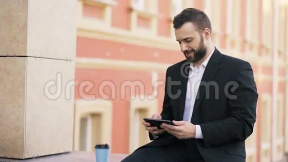在城市街道休息时年轻的大胡子商人在平板电脑上阅读新闻视频的预览图