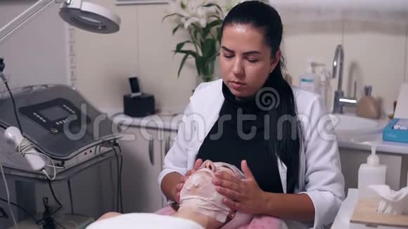 专业美容师在美容过程中用口罩给年轻女性做面部按摩视频的预览图