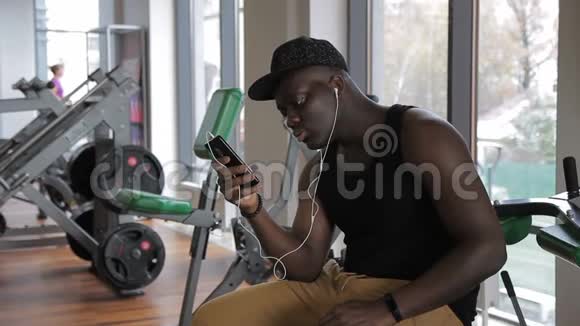 健身房里的非洲男人坐着通过手机耳机听音乐视频的预览图