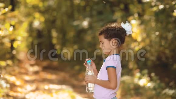 小女孩在秋天的树林里喝塑料瓶里的水秋天美丽的森林里的小女孩视频的预览图