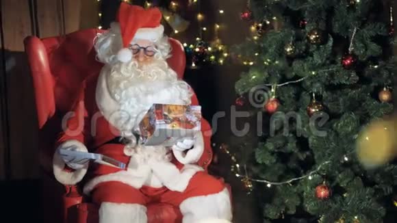 圣诞老人坐在圣诞树附近的椅子上包装一个礼盒视频的预览图