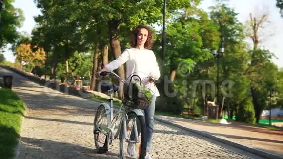 一个穿着白色t恤和蓝色牛仔裤的年轻女子手持她的城市自行车走在城市公园的鹅卵石路上视频的预览图