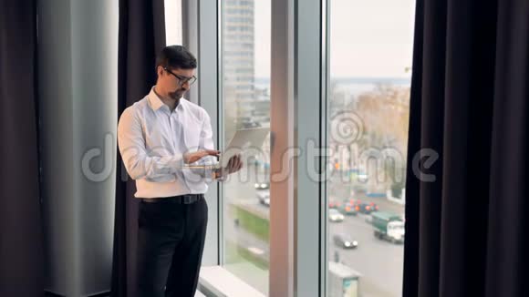 一个商人站在高高的窗户附近在他的笔记本电脑上工作视频的预览图