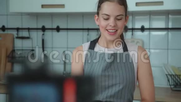 穿灰色围裙的年轻女子在灯光厨房录制视频视频的预览图