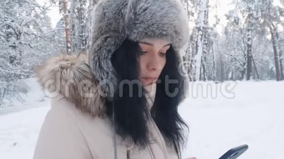冬天白雪皑皑的森林里一位戴着温暖帽子的白种人年轻女士用智能手机紧紧地抱在一起视频的预览图