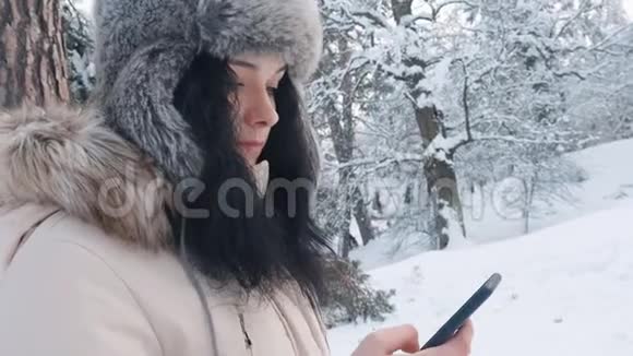 冬天白雪皑皑的森林里一位戴着温暖帽子的白种人年轻女士用智能手机紧紧地抱在一起视频的预览图