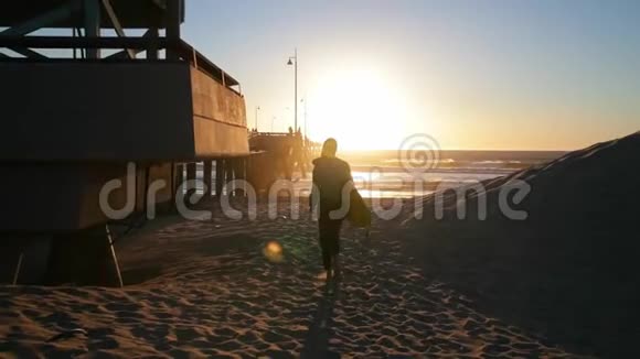 小女孩拿着冲浪板在海滩上带着海浪走向海洋的女人美丽的日落风吹沙子视频的预览图