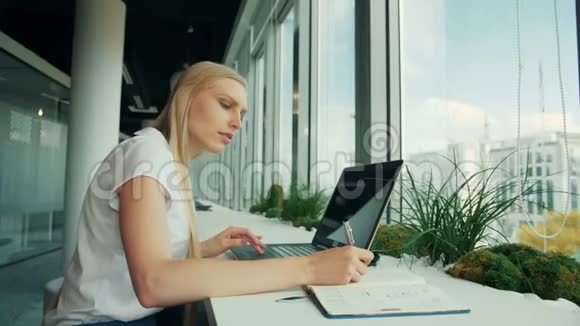 有笔记本电脑和文件的正式女人女商人一边坐在桌子旁一边用纸写字一边拿着笔记本电脑视频的预览图