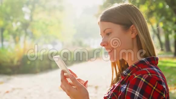 微笑的年轻女人享受大自然坐在绿色公园的长椅上用手机浏览视频的预览图