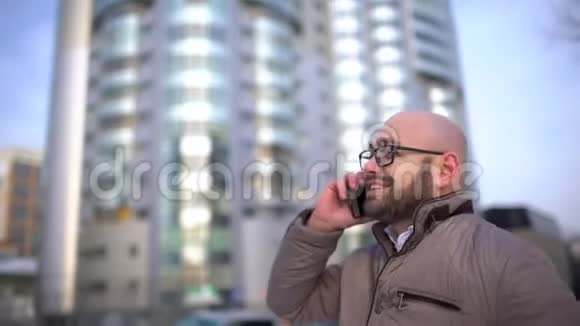 年轻的商人带着智能手机穿过城市街道那个年轻人拿着电话沿街走去他在旁边说话视频的预览图