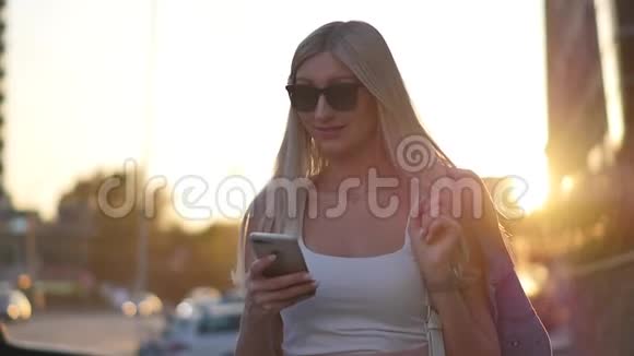 迷人的金发女郎在户外的智能手机上写信息一个女人用一个数字小工具滚动着社交视频的预览图