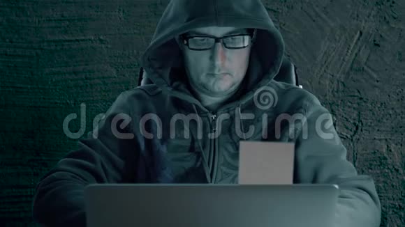 穿着帽衫和戴着眼镜的黑客在晚上坐在桌子旁并编码黑客把备忘录从视频的预览图