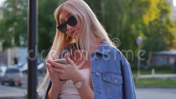 迷人的金发女郎在户外的智能手机上写信息一个女人用一个数字小工具滚动着社交视频的预览图