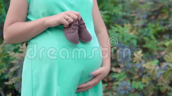 怀孕的女孩手里拿着绿色灌木丛背景下孩子们的鞋子视频的预览图