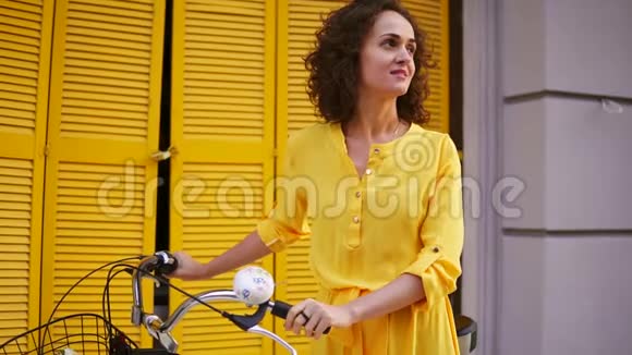 一个穿着黄色长裙的快乐黑发女人站在黄色的栅栏旁手里拿着她的城市自行车车把视频的预览图