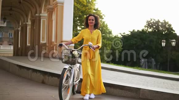 漂亮的快乐女人穿着一件黄色的长裙子走在城市里手里拿着她的城市自行车车把车把上有花视频的预览图