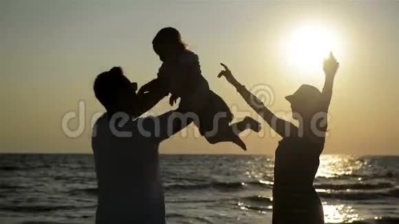 太阳落山时母亲在夏帽和父亲与小女儿在海边的黑暗剪影强壮的男人旋转视频的预览图