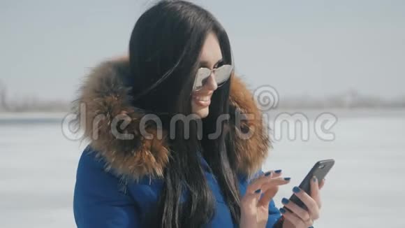 年轻美丽的黑发女人戴着太阳镜在冬天下雪的背景下使用智能手机的肖像视频的预览图