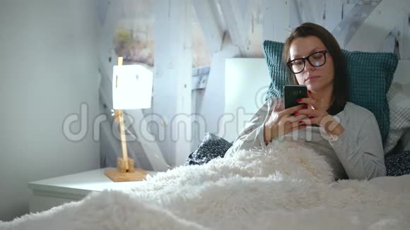 戴眼镜的女人躺在床上睡觉前使用智能手机视频的预览图