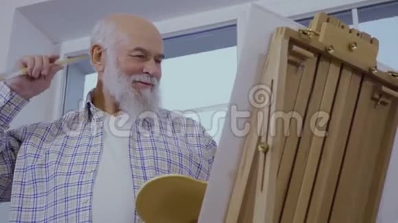 白发苍苍的老人在靠近窗户的画架上画画视频的预览图
