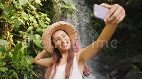 穿着白色连衣裙和草帽的年轻快乐混合种族旅游女孩用手机拍摄令人惊叹的野生丛林视频的预览图