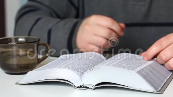一个年轻人坐在桌子旁拿着一块银片读圣经他从杯子里喝水视频的预览图