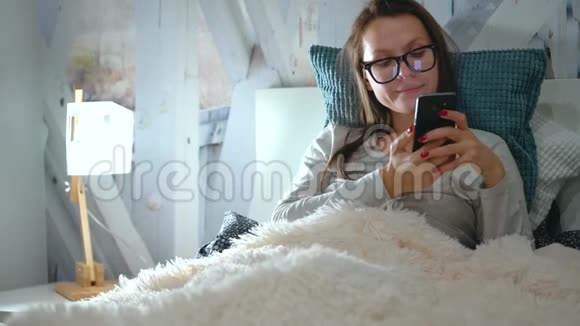 戴眼镜的女人躺在床上睡觉前使用智能手机视频的预览图