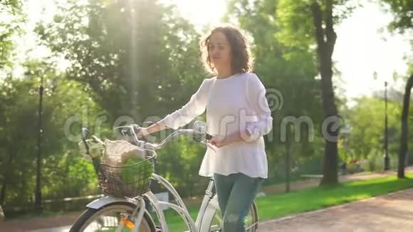 穿着白色衬衫和蓝色牛仔裤的漂亮女人拿着她的城市自行车走在城市公园的小巷视频的预览图