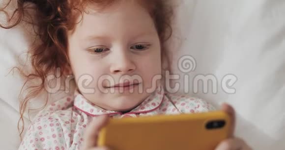 有趣的红发小女孩躺在床上饶有兴致地在现代黄色智能手机上观看视频视频的预览图