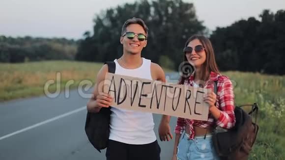 年轻的旅行夫妇相爱站在路上举着一个标志冒险快乐的微笑的朋友们正在注视视频的预览图